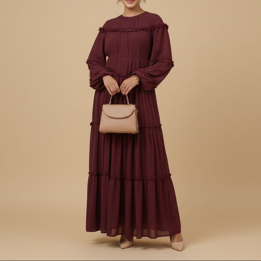 Woman wearing a burgundy long dress standing in a softly lit room with a white chair and decorative elements.