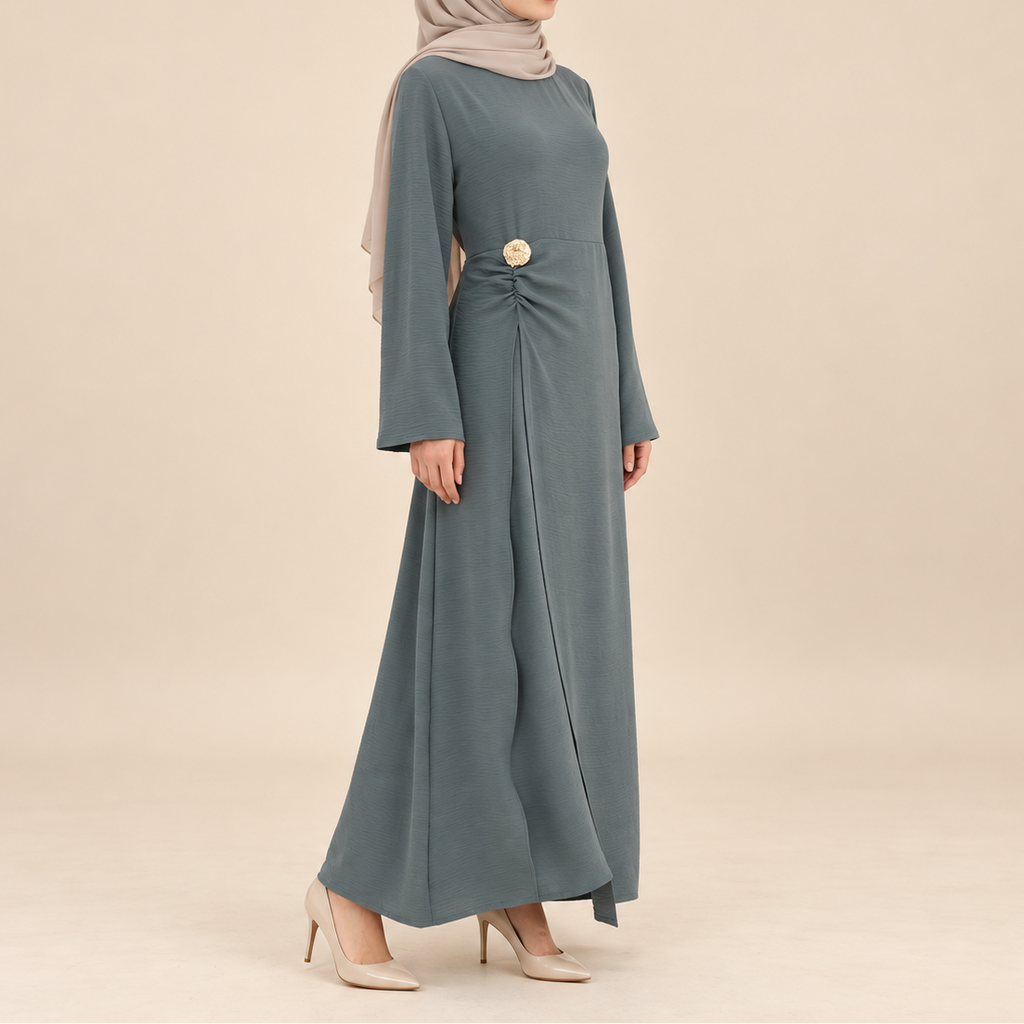 A woman modeling a long-sleeved, floor-length modest dress in a muted blue-gray color, styled with a light taupe hijab and beige heels against a solid beige studio background.