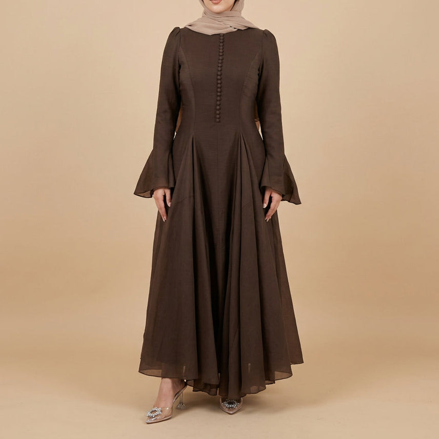 A woman modeling a long-sleeved, dark brown maxi dress with a buttoned bodice and flared bell sleeves, standing against a plain white studio background.