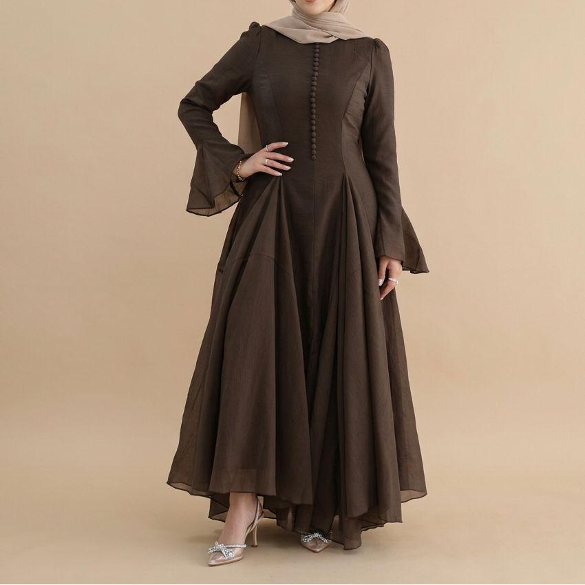 A woman modeling a long-sleeved dark brown modest maxi dress with a buttoned front and flared bell sleeves, paired with a light beige hijab, against a solid beige studio background.