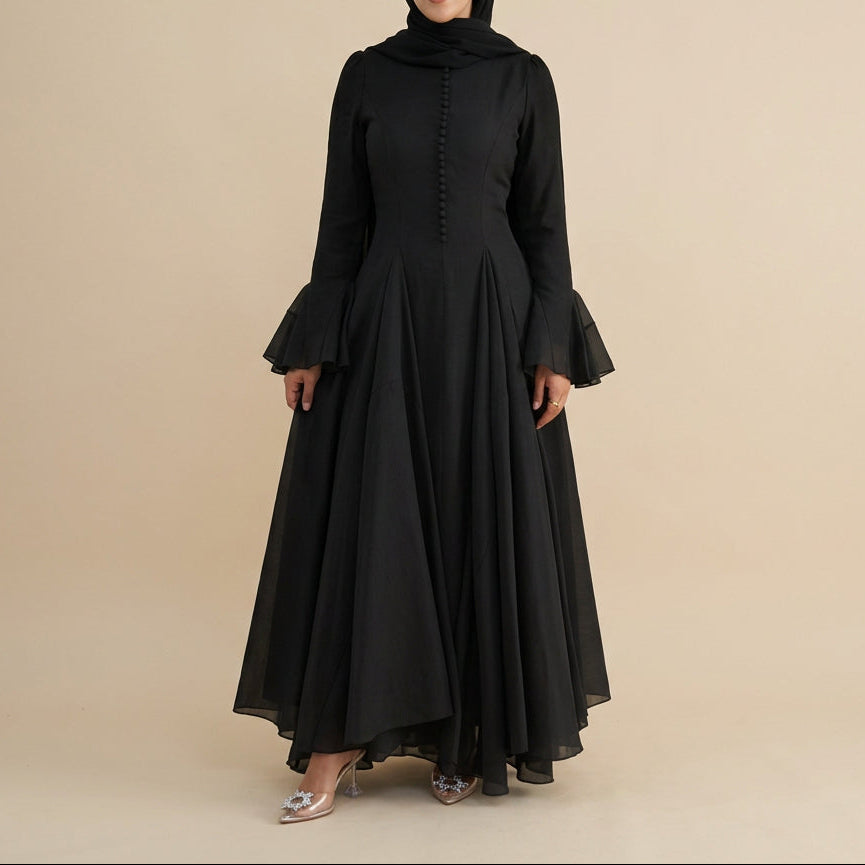 A woman modeling a long black dress with a high mock neck, buttoned front, and dramatic flared bell sleeves, photographed against a plain light gray studio background.