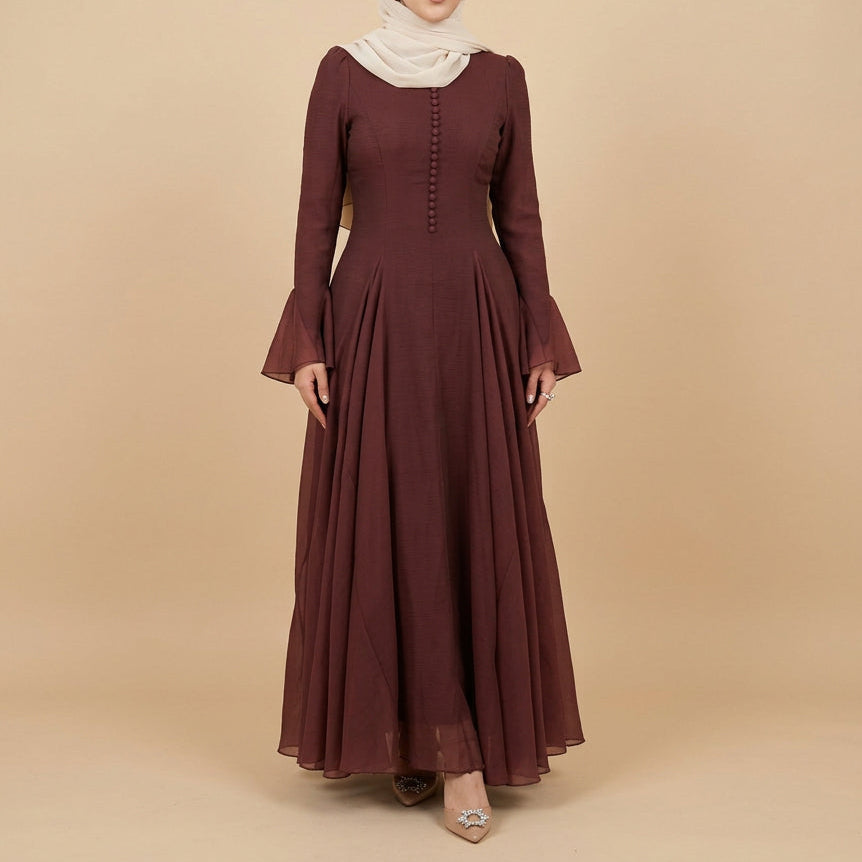 A woman modeling a long-sleeved, floor-length maroon dress with a gathered waist and buttoned bodice, set against a plain light gray studio background.
