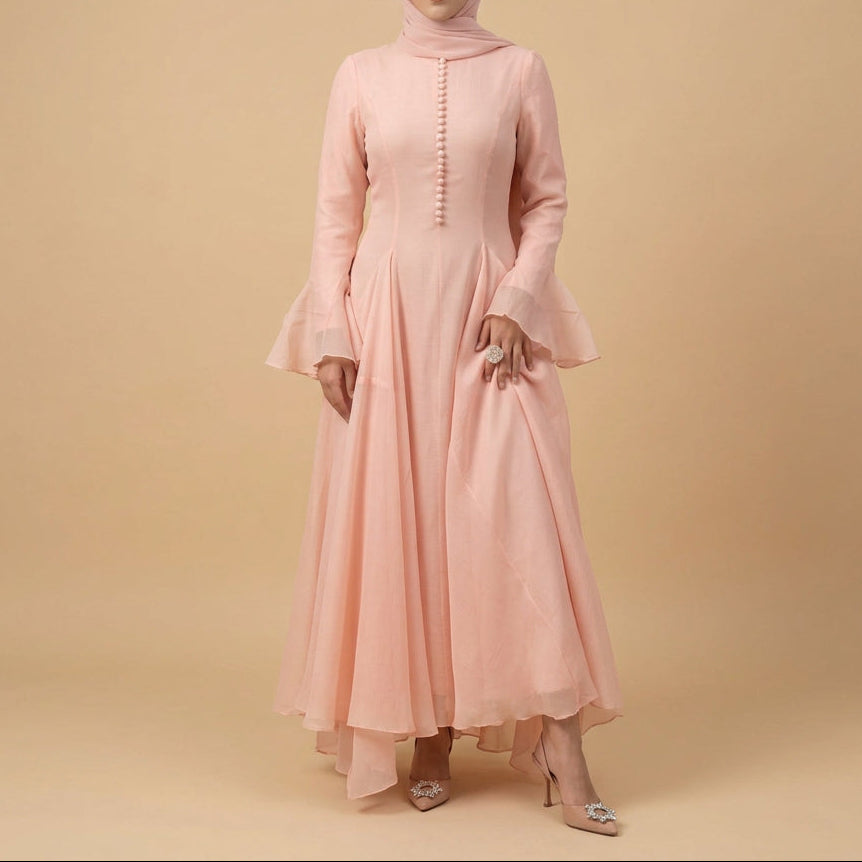 A woman modeling a long, flowing pink gown with a sheer draped overlay and flared sleeves, standing against a plain light gray studio background.