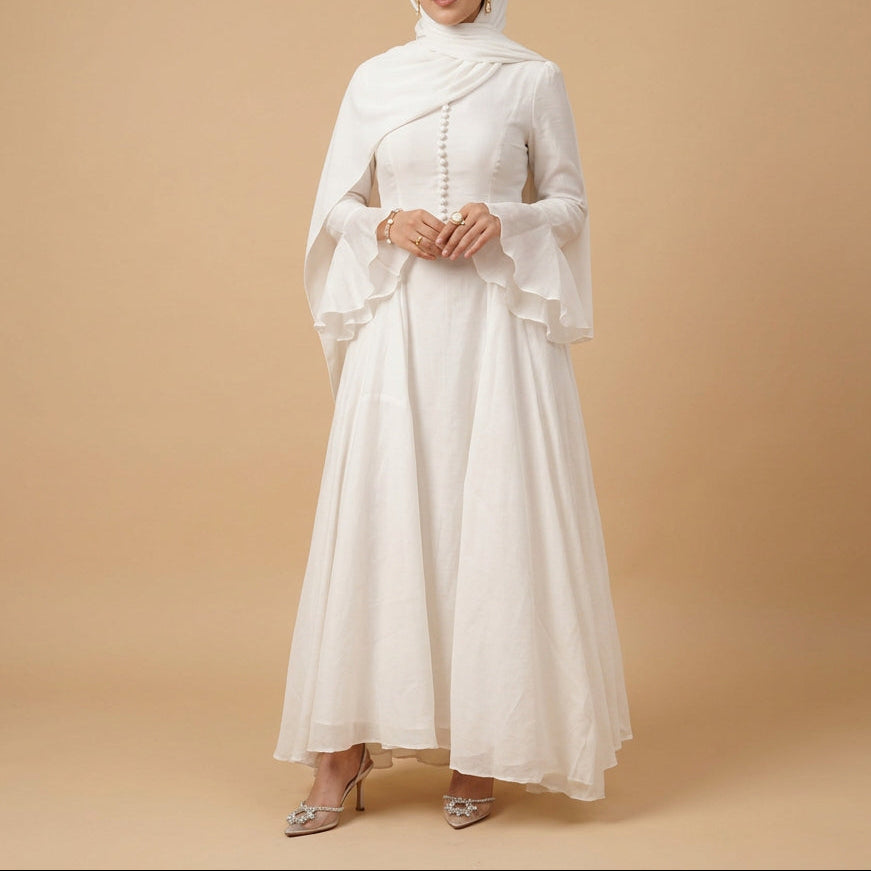 A woman wearing a flowing white dress with a sheer draped overlay and long ruffled sleeves, standing against a plain light background.