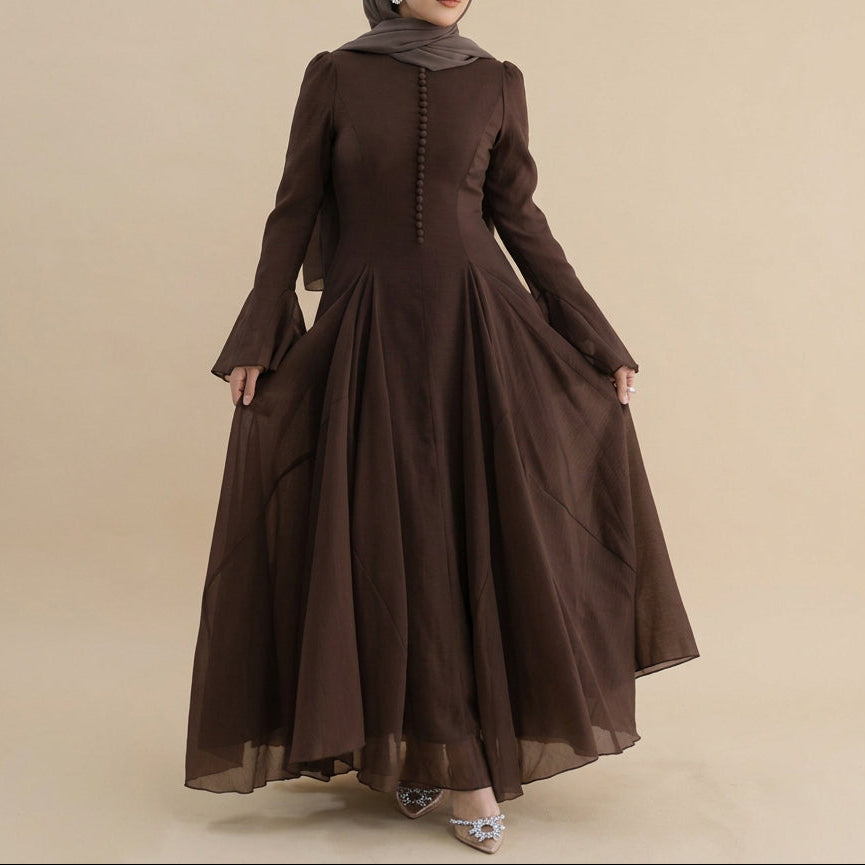 A woman modeling a long-sleeved, floor-length brown dress with a gathered waist and buttoned front, photographed against a plain light gray studio background.