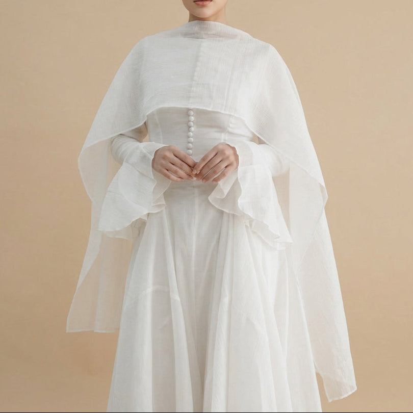 A woman wearing a flowing white dress with a sheer draped capelet and long ruffled sleeves, standing against a plain light gray studio background.