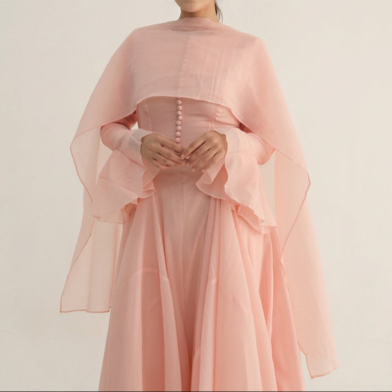 A woman wearing a light pink traditional dress with a matching sheer draped overlay, standing against a plain light gray studio background.