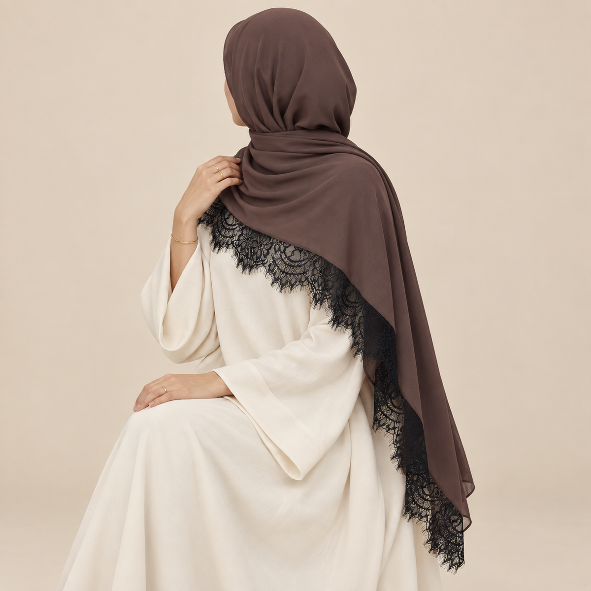 A woman wearing a brown hijab with black lace trim, shown from the back against a plain beige studio background.