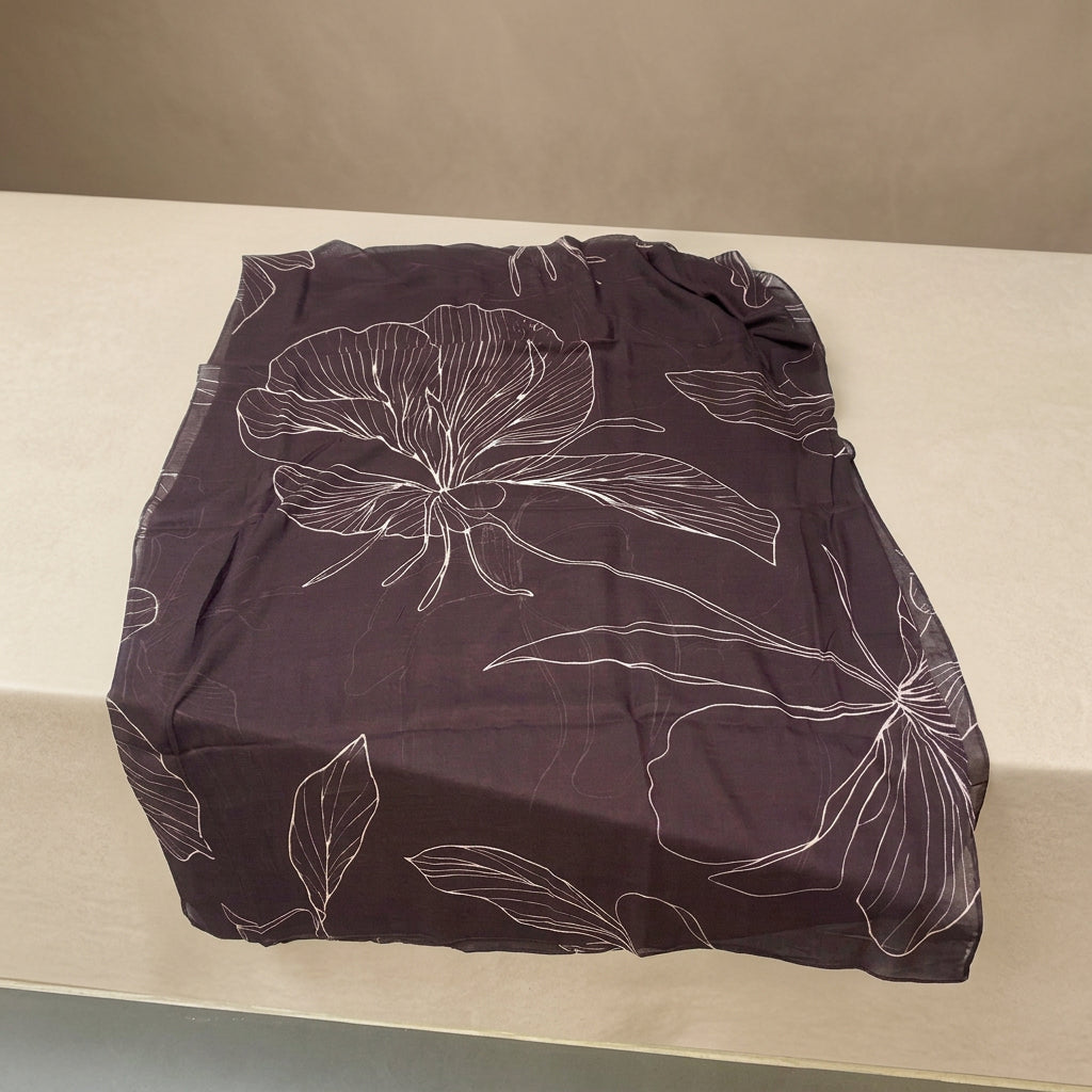 A dark brown sheer scarf with a white floral line-art pattern, laid flat on a white table with decorative flowers in the background.