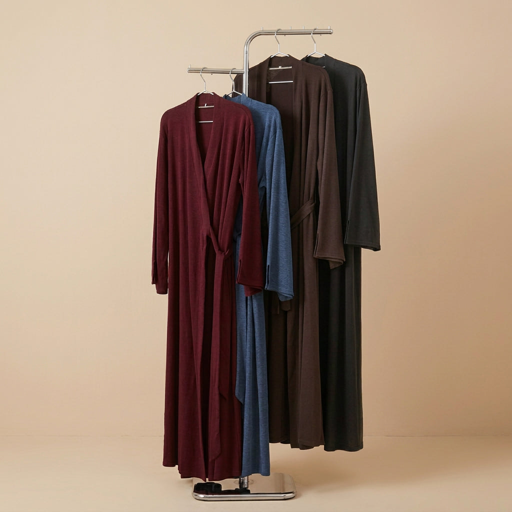 Three long draped robes or kaftan-style garments hanging on a metal rack against a white paneled wall. The front garment is burgundy, the middle is blue, and the back is dark brown or very dark taupe, with a green plant leaf visible at the right edge.