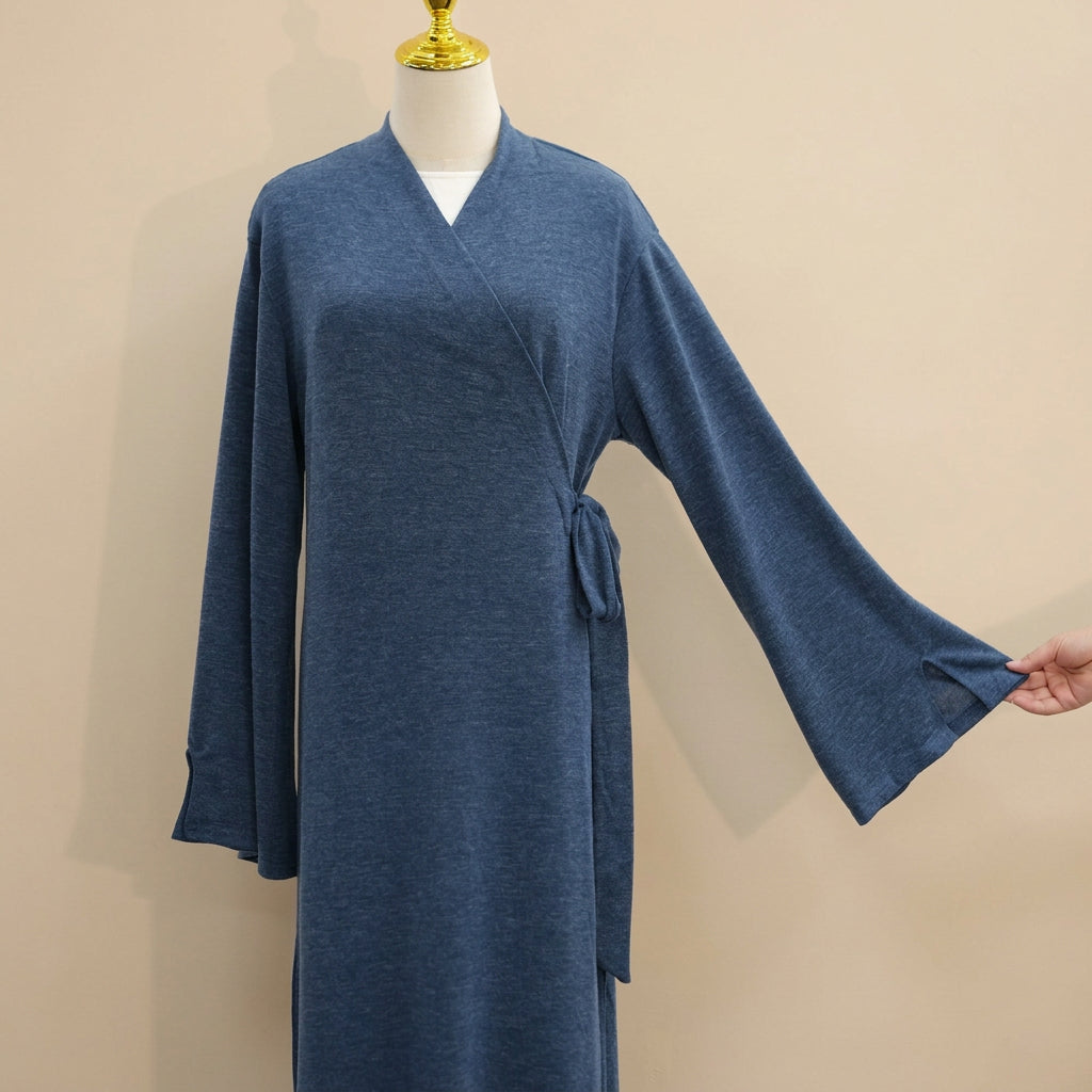 A slate blue wrap robe displayed on a mannequin torso with a gold neck cap, set against a white paneled wall. A hand is shown stretching the wide sleeve of the robe.