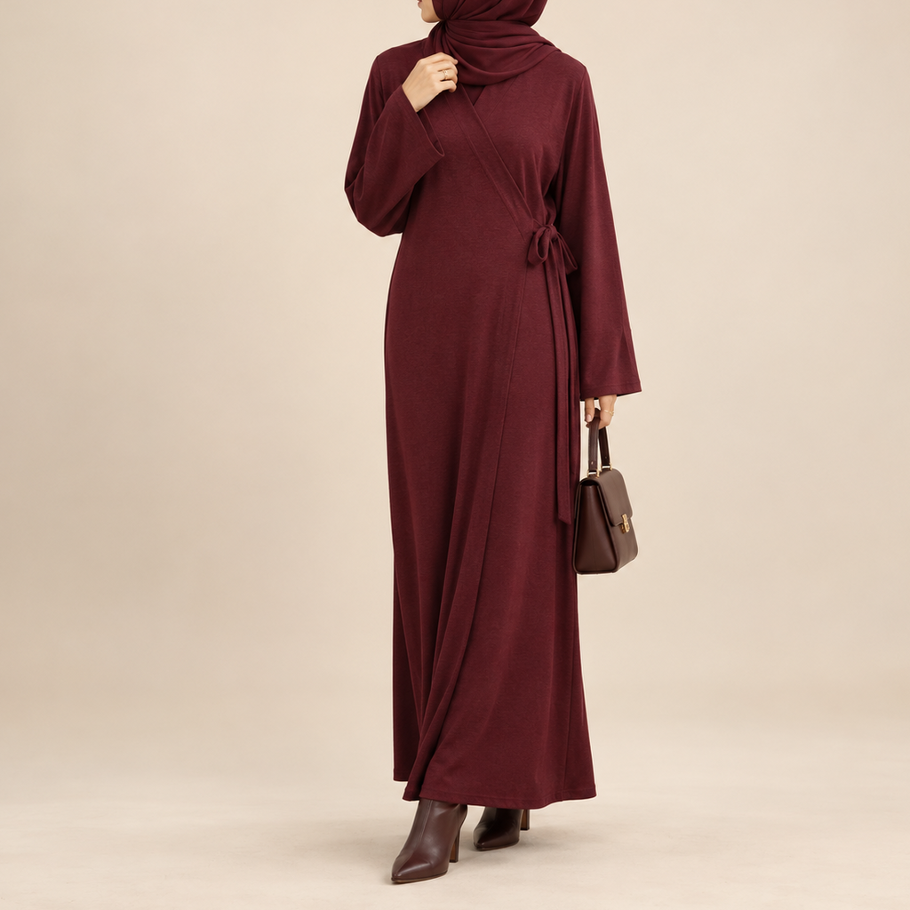 A woman wearing a long burgundy maxi abaya dress with a matching hijab, shown from the back against a plain beige studio background. She is holding a small brown handbag and wearing dark brown ankle boots.