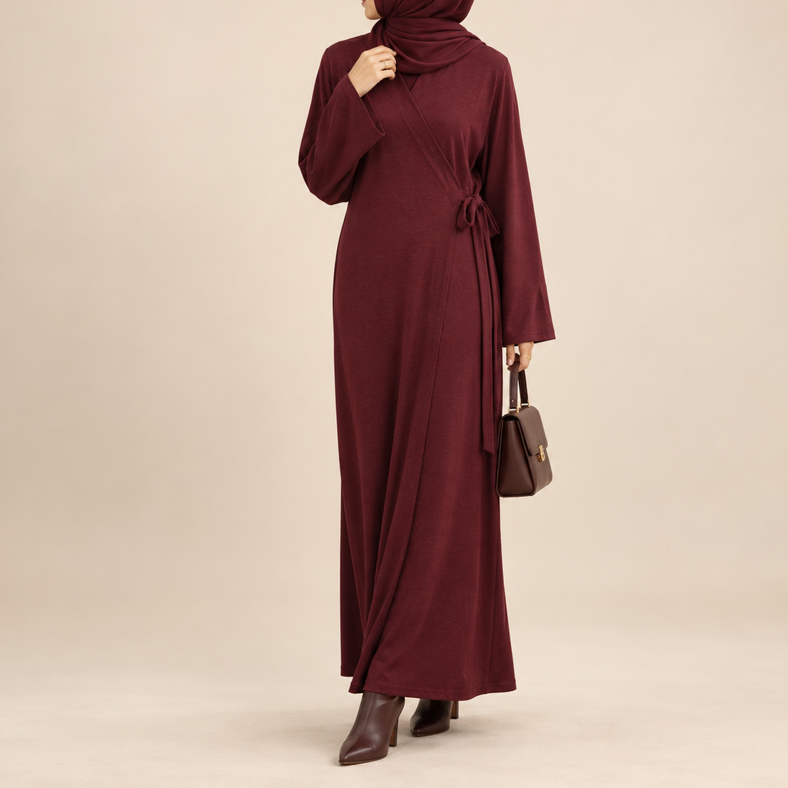 A woman wearing a long burgundy maxi abaya dress with a matching hijab, shown from the back against a plain beige studio background. She is holding a small brown handbag and wearing dark brown ankle boots.