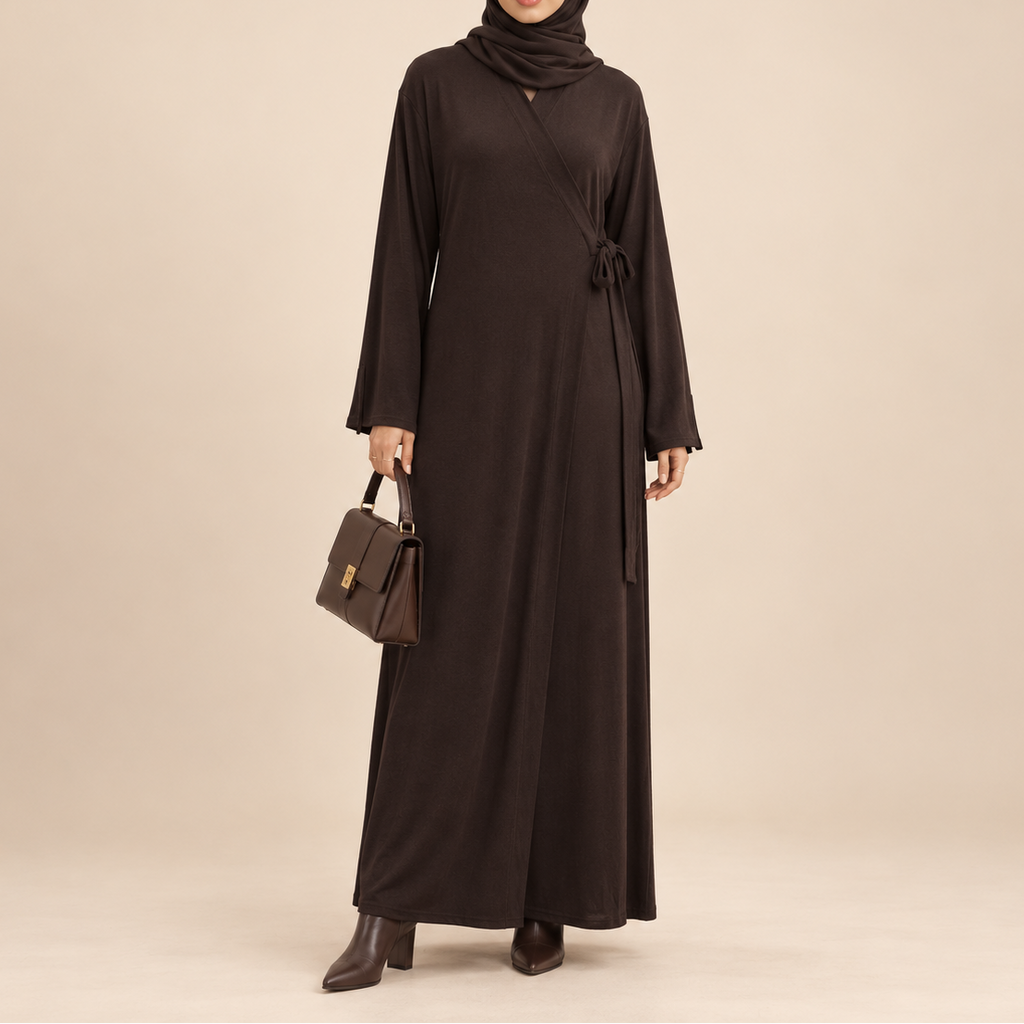 A woman modeling a long-sleeved, dark brown modest maxi dress with a matching hijab and carrying a small brown handbag, set against a solid beige studio background.