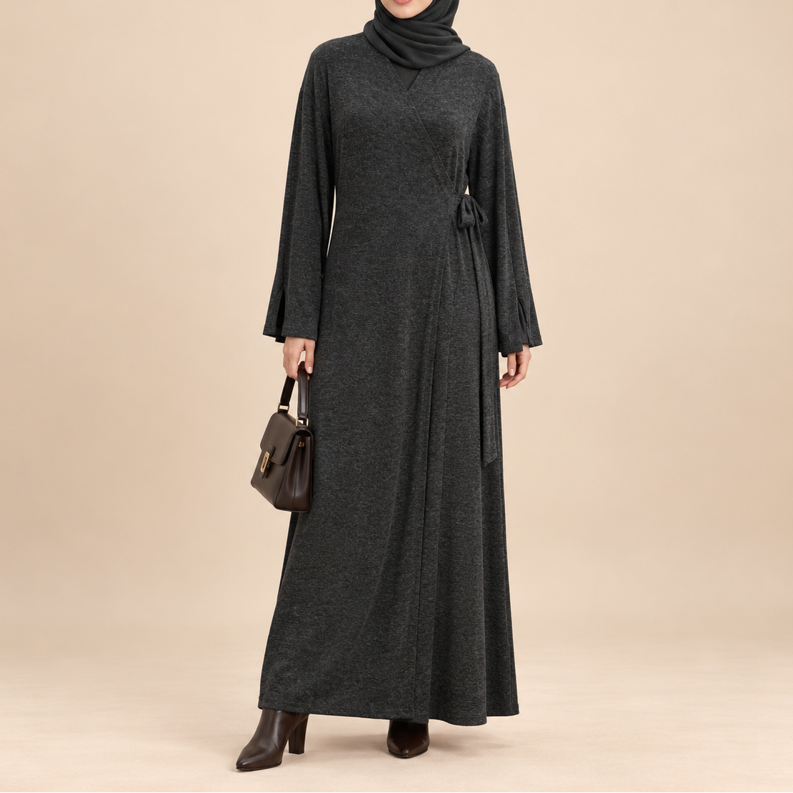 A woman modeling a dark charcoal gray long-sleeve maxi dress with a self-tie waist, paired with a matching hijab, dark brown handbag, and dark brown boots, set against a solid beige studio background.