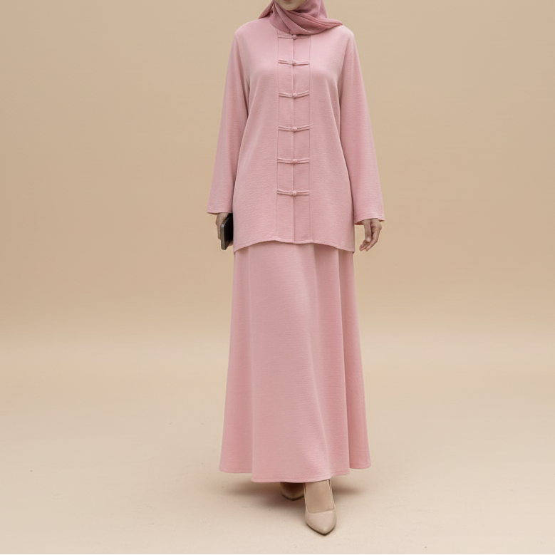 A woman wearing a light pink two-piece modest outfit with a matching hijab and carrying a small pink handbag, standing on a sidewalk in front of a beige building with large glass doors.