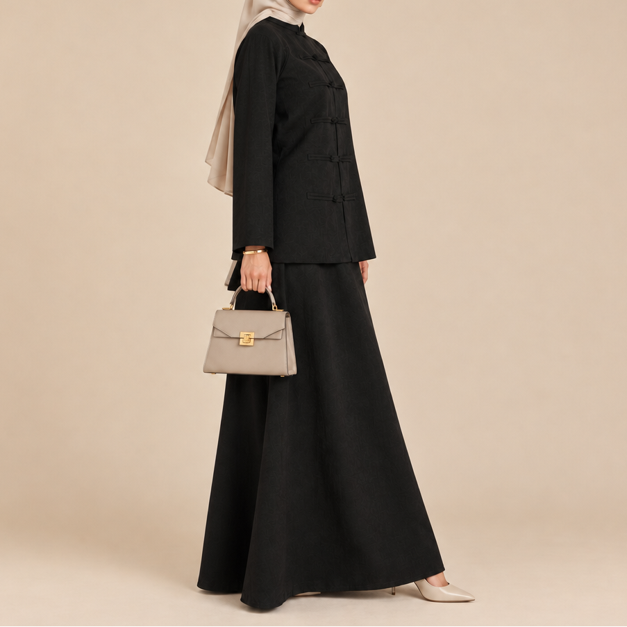A woman modeling a black two-piece modest outfit consisting of a long-sleeve button-front jacket and a matching maxi skirt, paired with a light beige hijab and holding a structured taupe handbag against a plain beige studio background.