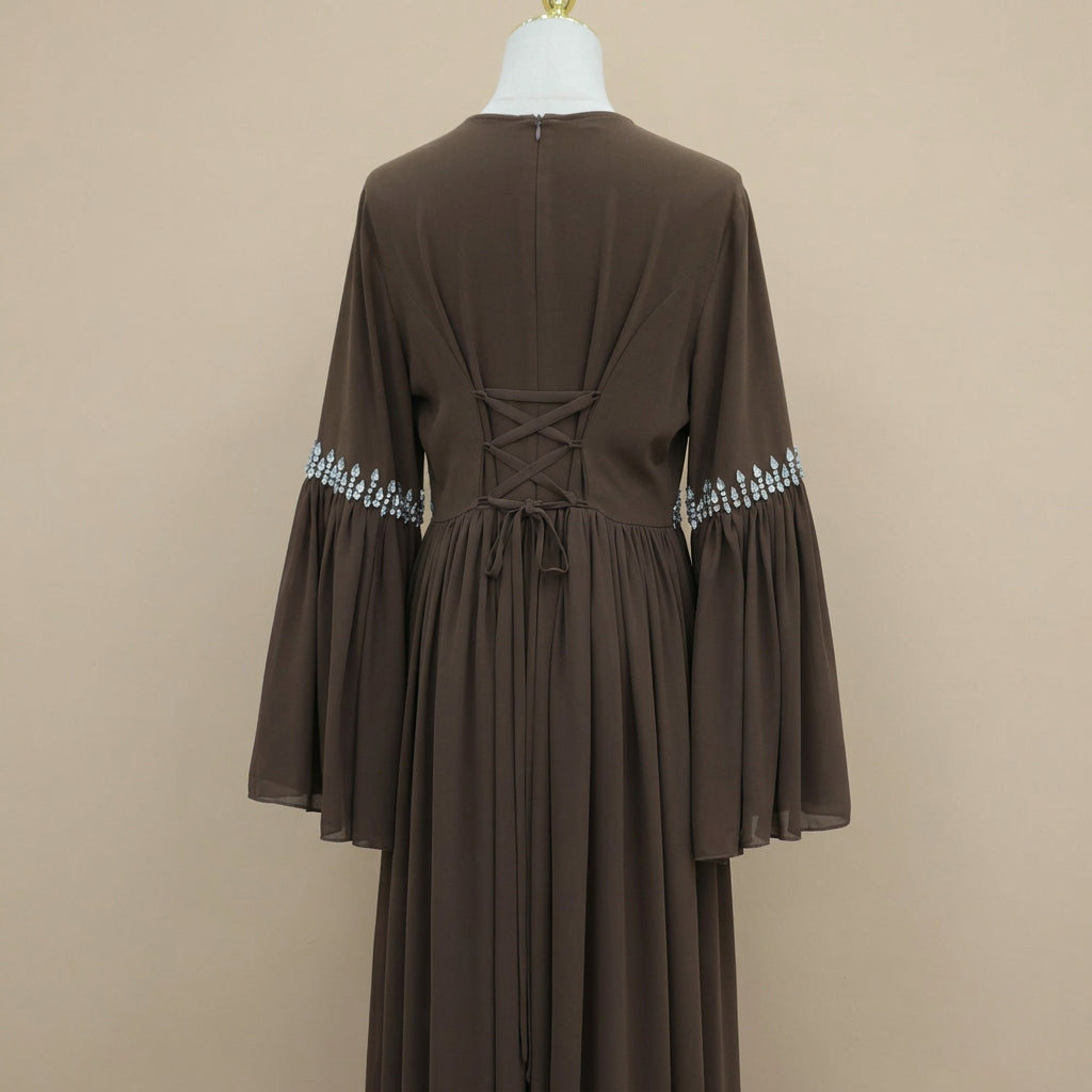 A brown long-sleeve maxi dress displayed on a mannequin from the back, featuring a lace-up back and silver beaded cuffs, set against a white paneled wall.