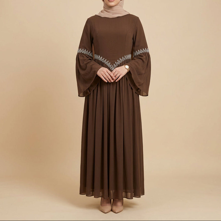 A brown long-sleeve maxi dress with a V-shaped silver embroidered pattern across the chest, displayed on a mannequin in front of a white paneled wall.