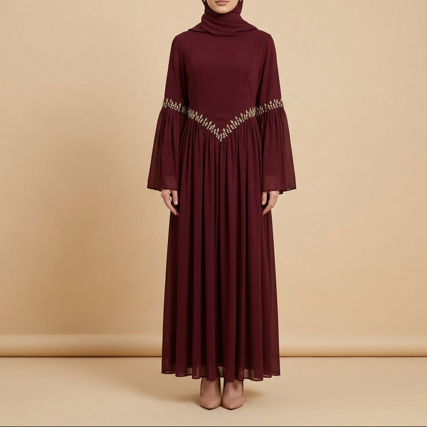A long-sleeved maxi dress in deep burgundy, displayed on a mannequin. The dress features a V-shaped neckline embellished with silver rhinestone beads and wide flared bell sleeves, set against a white paneled wall background.