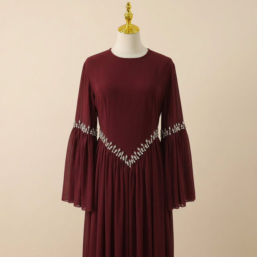 A long-sleeved burgundy maxi dress with a V-shaped embellished neckline and matching embellished cuffs, displayed on a mannequin against a white paneled wall.