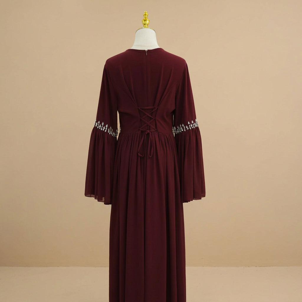 A long burgundy dress displayed on a cream dress form against a white paneled wall. The back view shows a V-neckline, crisscross lace-up back detail, a drawstring waist, and silver beaded trim on the flared sleeves.