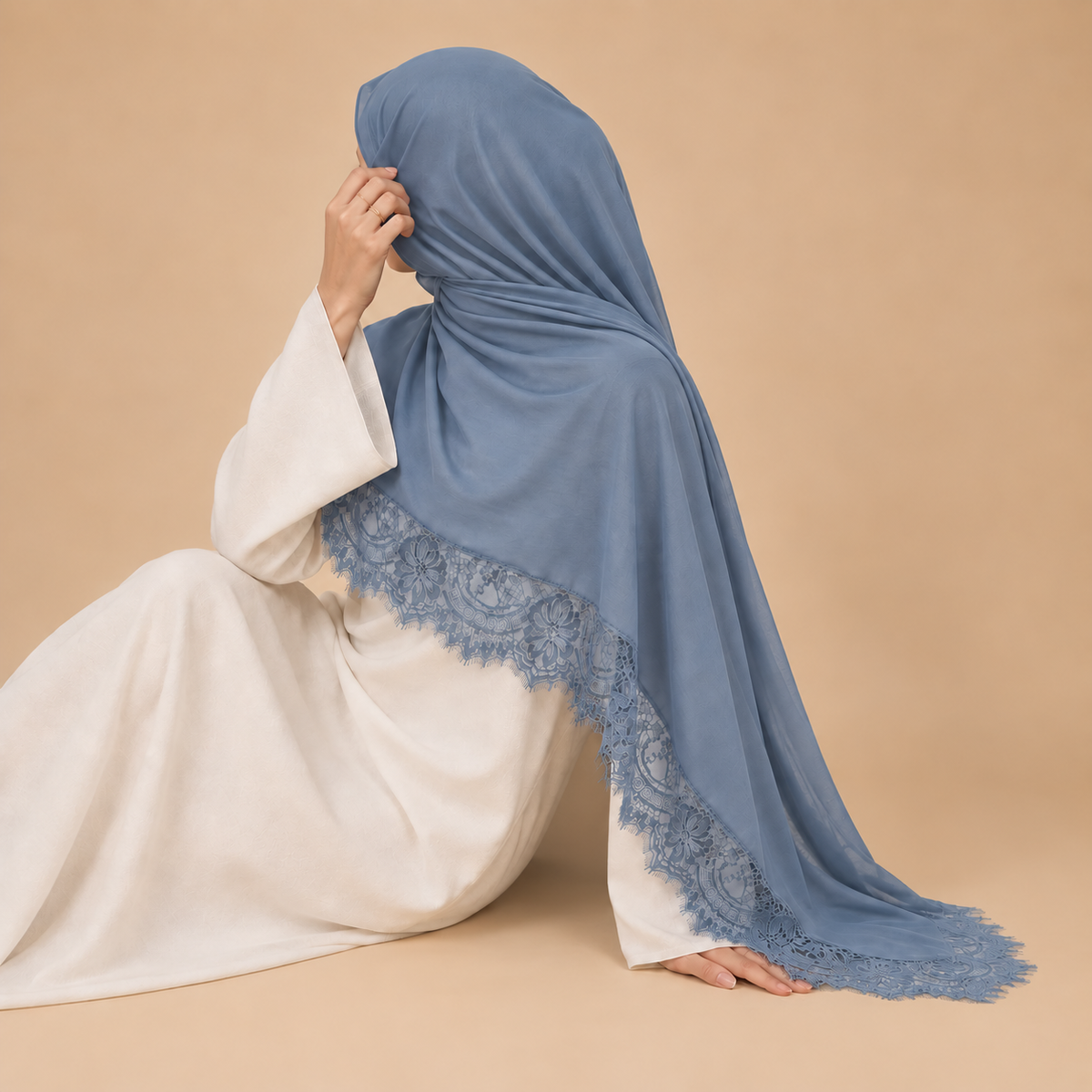 A woman wearing a light blue modal hijab with a lace-trimmed border, draped over her head and shoulders, posed against a solid beige studio background.
