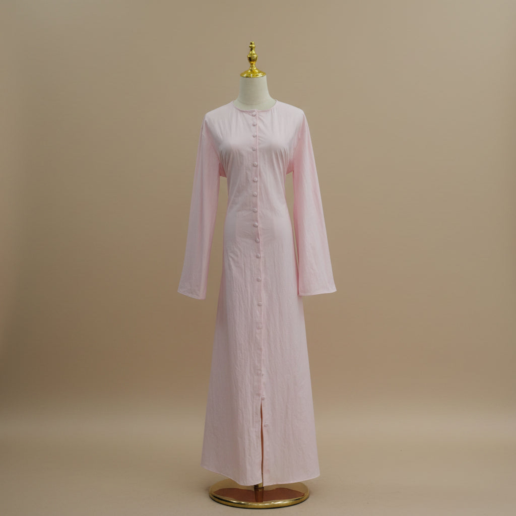 A long-sleeved light pink maxi dress displayed on a cream dress form with a gold finial and base, set against a white paneled wall.