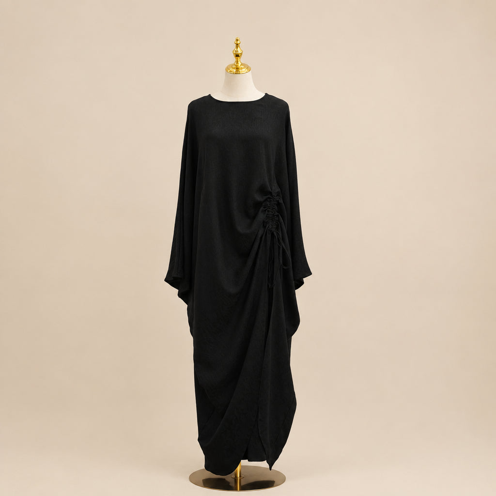 A black long-sleeve maxi dress displayed on a mannequin against a white paneled wall, with a small green plant leaf visible on the right side.