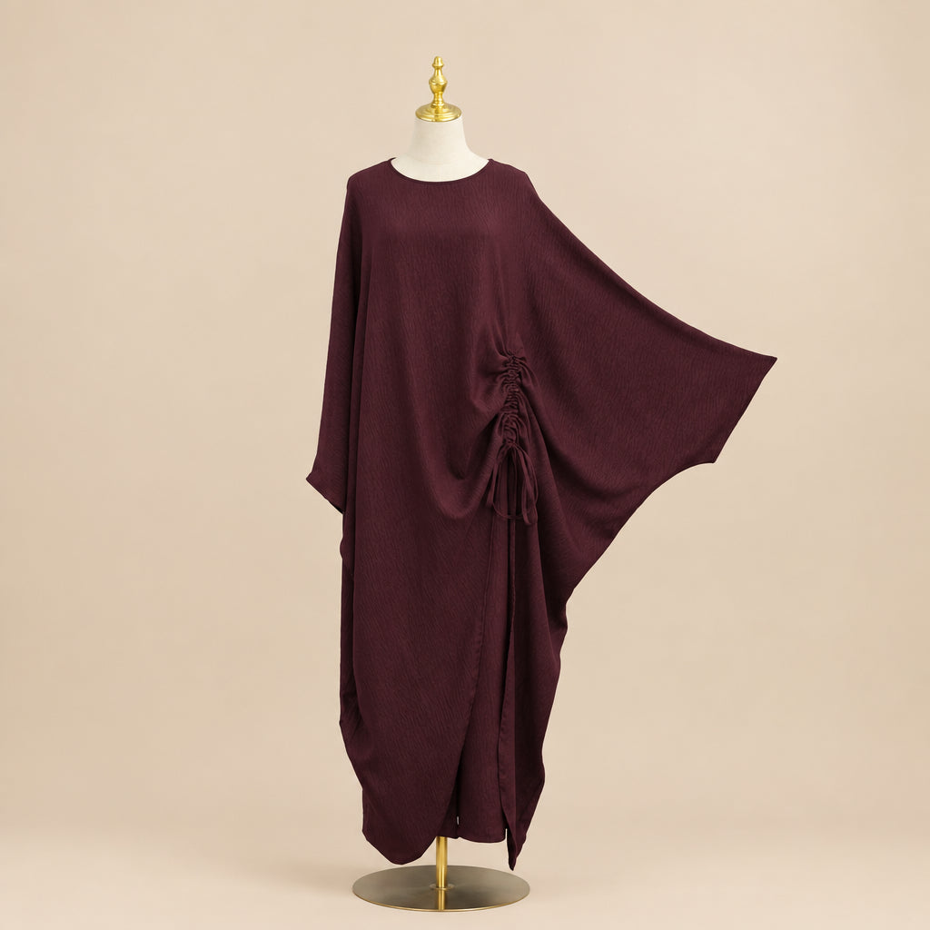 A dark burgundy batwing caftan dress displayed on a mannequin, with a hand holding out one of the wide sleeves to show its drape. The background is a white paneled wall with a green plant leaf visible on the right.