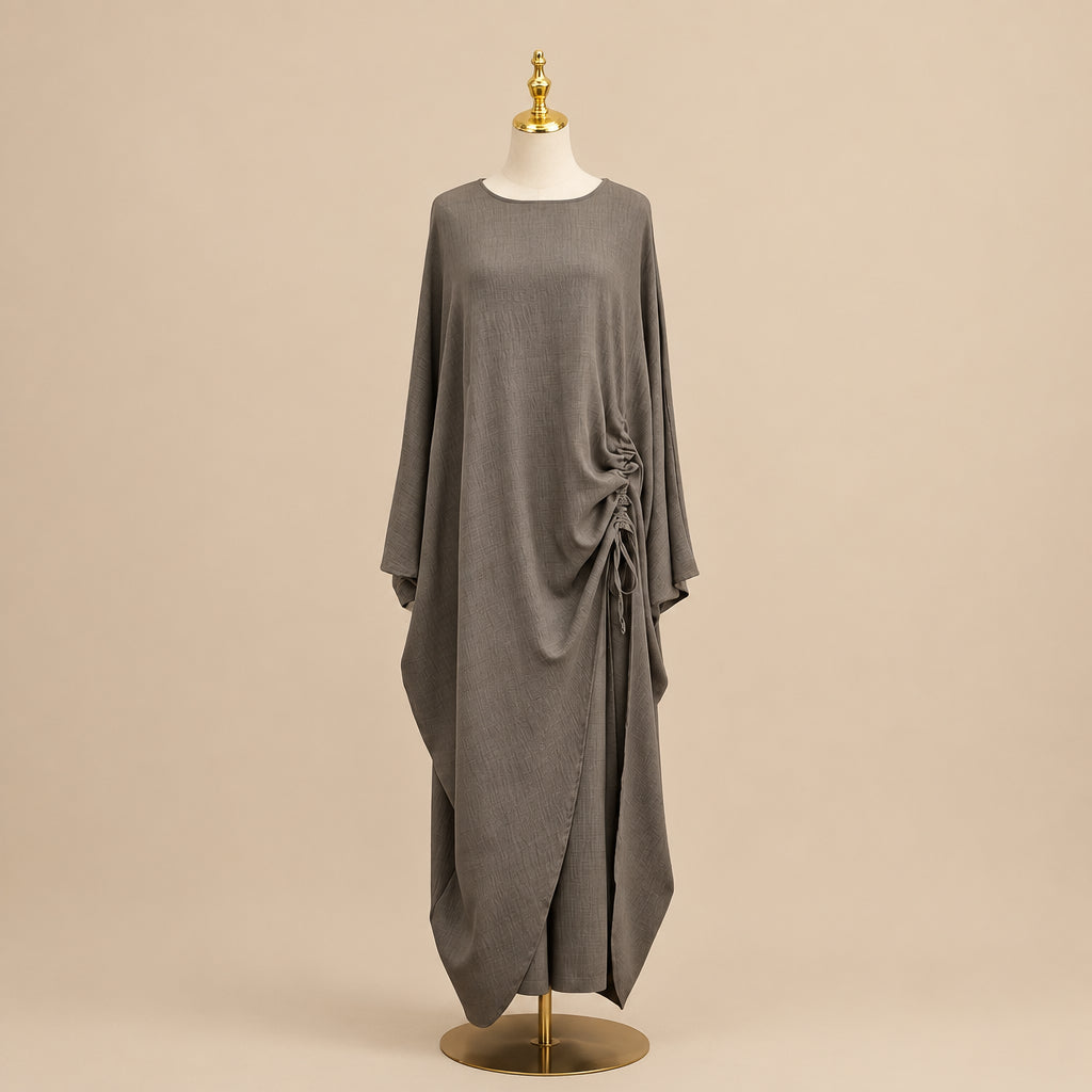 A taupe-brown long-sleeve maxi dress with an asymmetrical draped side detail, displayed on a mannequin in front of a white paneled wall.