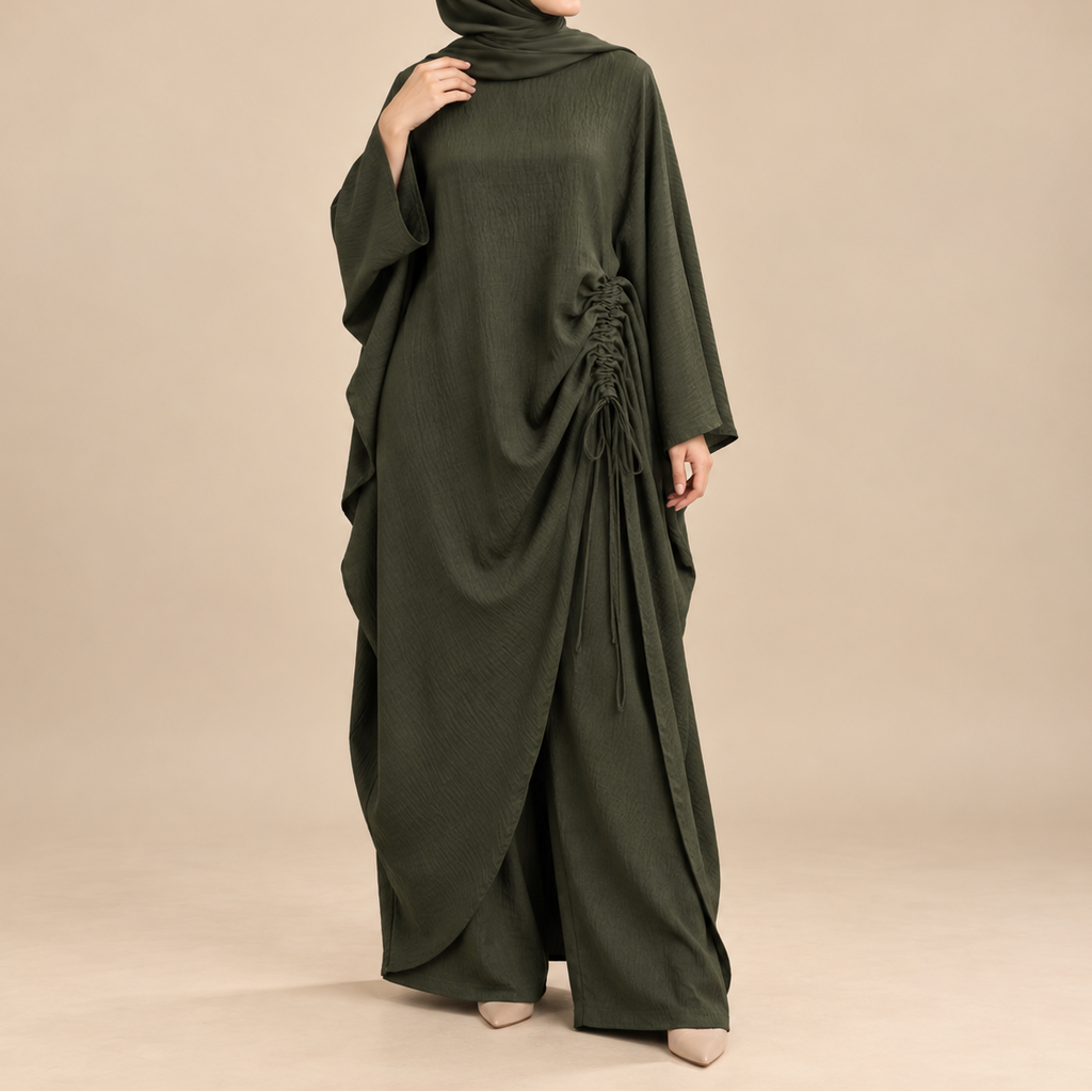 A woman modeling a dark olive green modest outfit consisting of a long-sleeved draped abaya with side lace-up detailing and a matching hijab, set against a solid beige studio background.