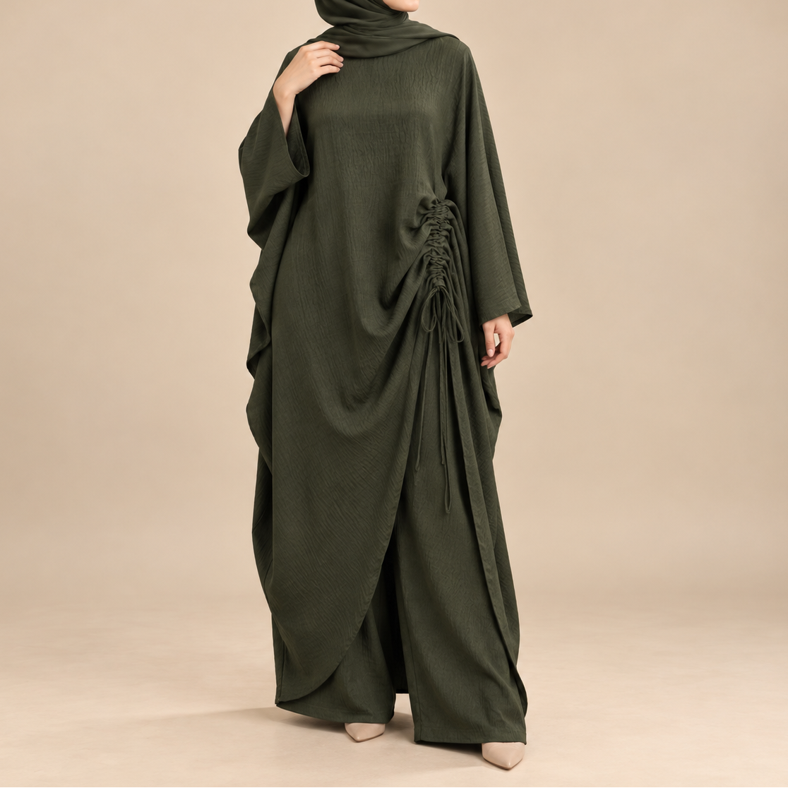A woman modeling a dark olive green modest outfit consisting of a long-sleeved draped abaya with side lace-up detailing and a matching hijab, set against a solid beige studio background.