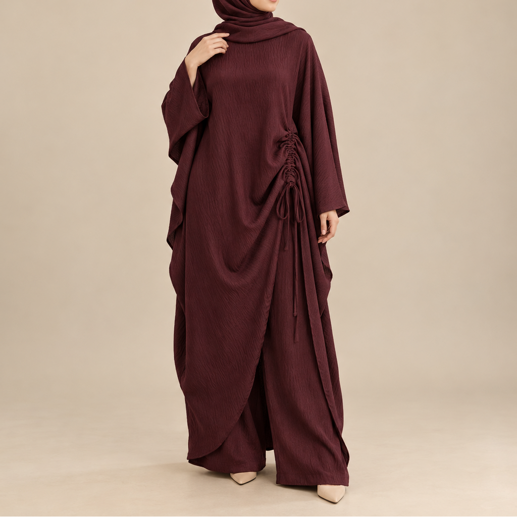 A woman modeling a deep burgundy hijab and matching draped abaya with side lacing, photographed against a plain beige studio background.
