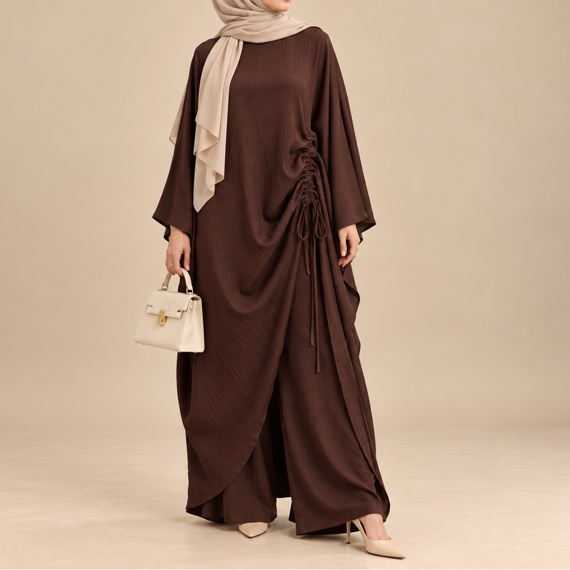 A woman modeling a dark brown modest dress with a side drawstring, a light beige hijab, and a small cream handbag against a solid beige studio background.