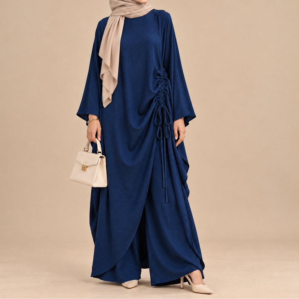 A woman modeling a navy blue modest maxi dress with a draped side detail and drawstring, paired with a beige hijab, holding a small cream handbag against a solid beige studio background.
