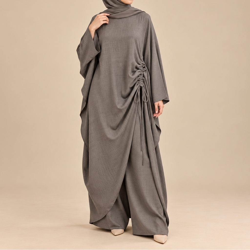 A woman wearing a draped, floor-length gray abaya with a matching hijab, standing against a plain beige studio background.