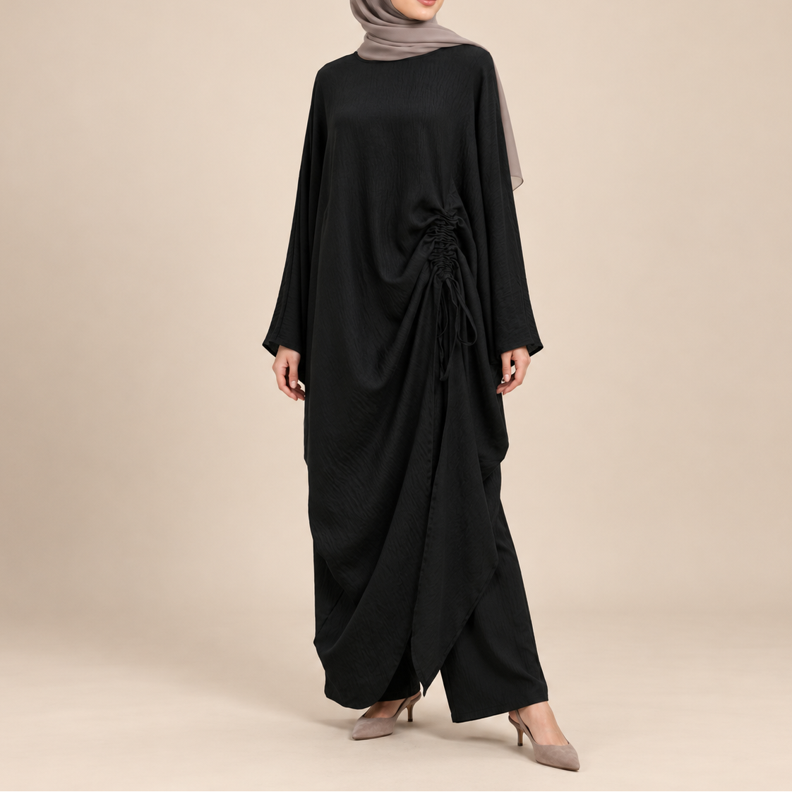 A woman modeling a black long-sleeved abaya dress with a draped, gathered design and a matching taupe-gray hijab, standing against a solid beige studio background.
