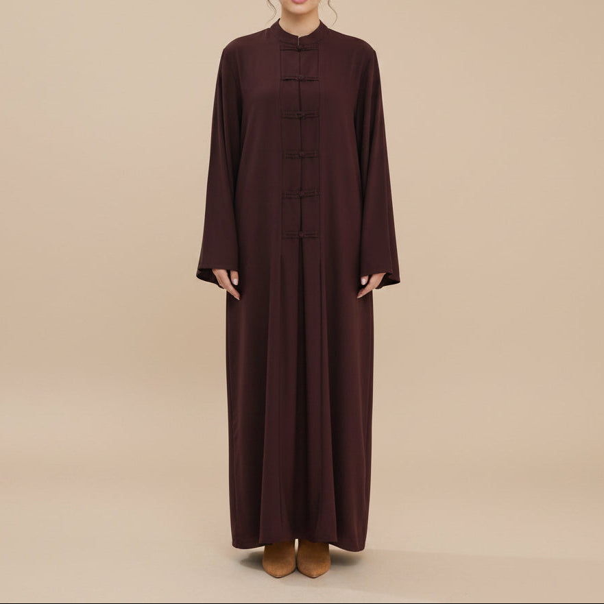 A woman stands against a plain light gray studio background wearing a long-sleeved abaya, ankle-length dark brown dress with traditional Chinese-style frog closures down the front.
