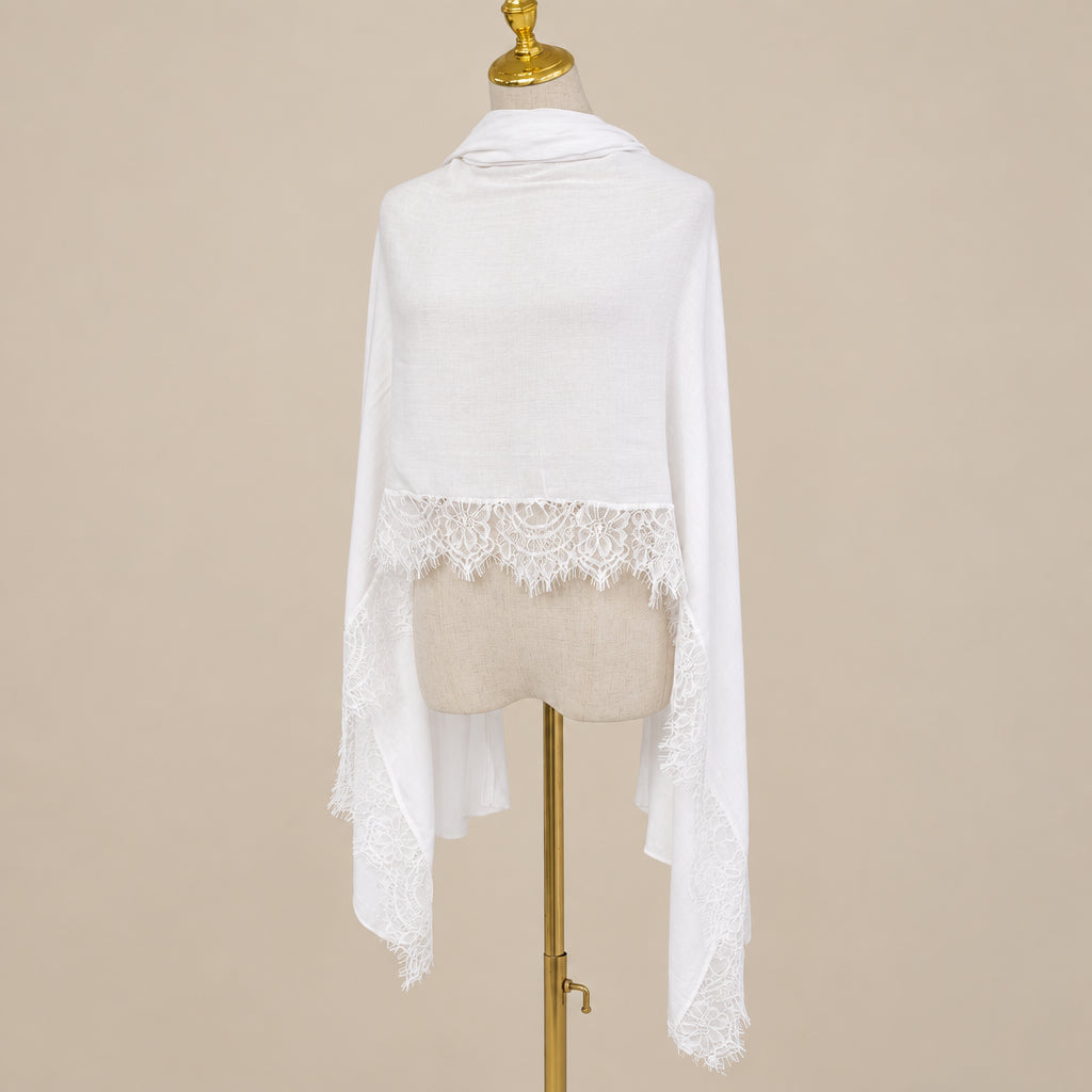 A white lightweight shawl with delicate lace trim along the hem, draped over a beige mannequin torso on a gold stand against a light gray wall.