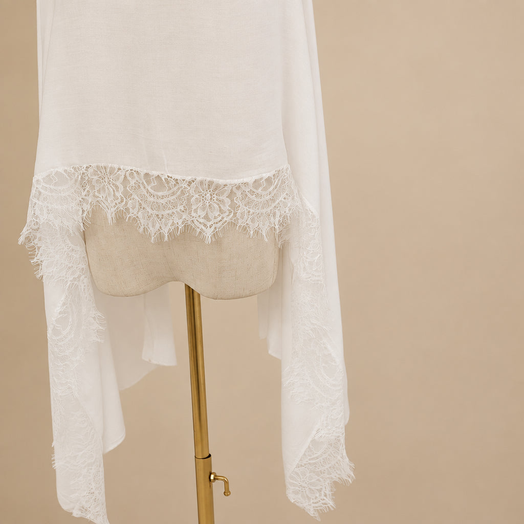 A long white sheer fabric with delicate floral lace trim draped over a beige fabric mannequin torso on a gold stand, set against a plain light gray background.