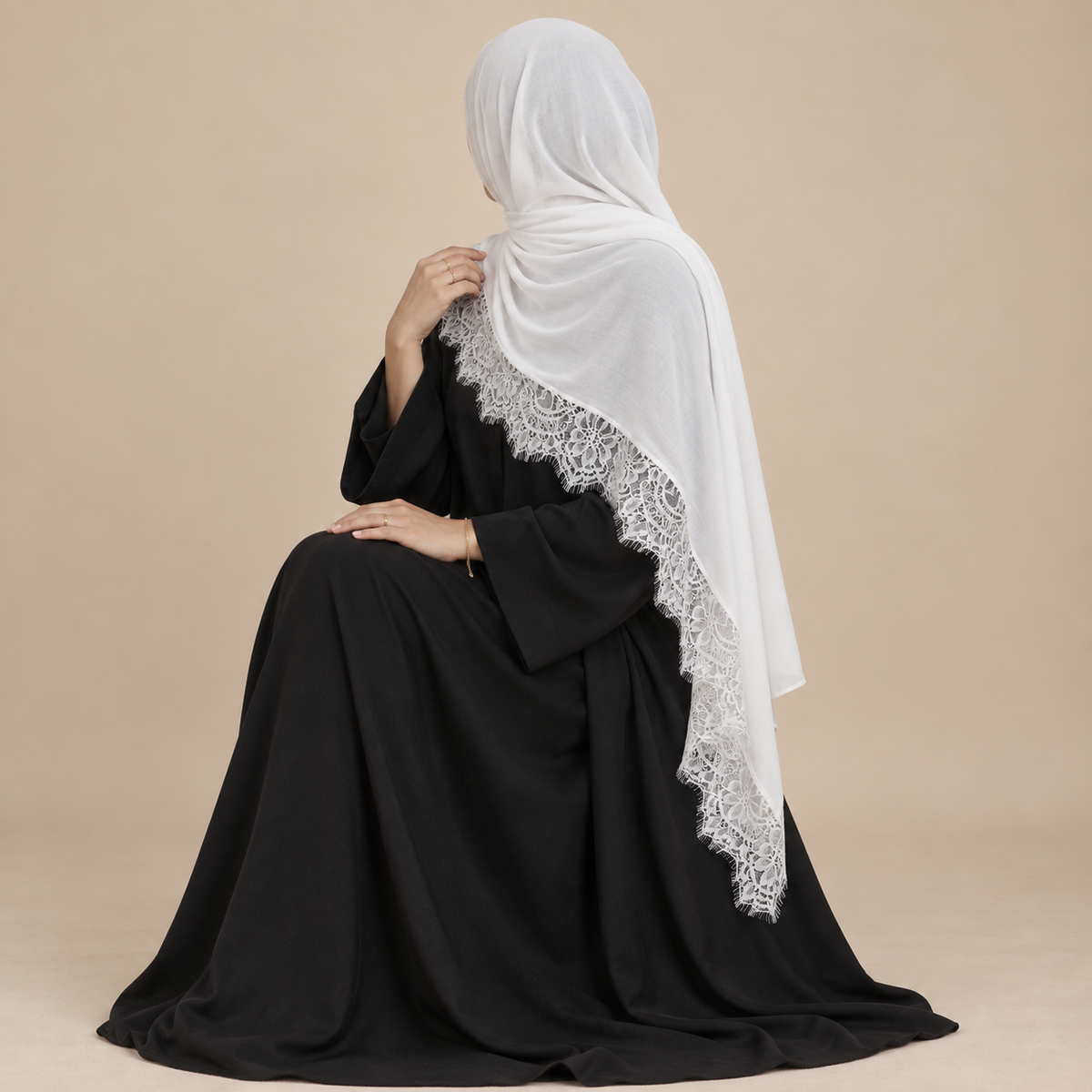 A woman wearing a black abaya and a white hijab draped over her shoulders, photographed from the back against a plain beige studio background.