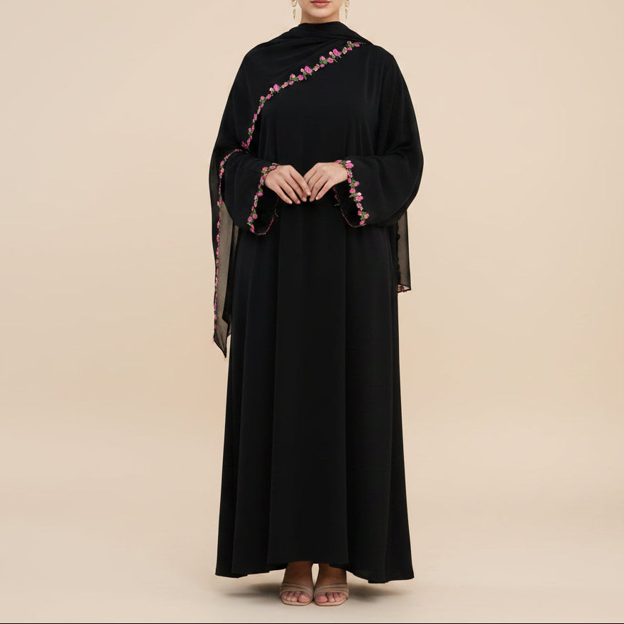 A woman modeling a long black abaya dress with wide sleeves and pink floral embroidery along the neckline and sleeve cuffs, standing against a plain white studio background.