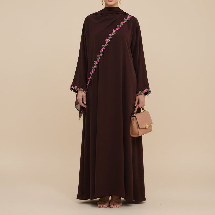A woman modeling a long, dark brown modest dress with pink floral embroidery along the shoulder and sleeve cuffs, standing against a white studio background.