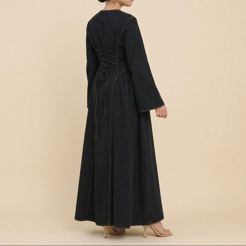 A woman wearing a long black wide-sleeved dress with a lace-up back, standing in profile against a white studio background with a decorative pampas grass plant and white cushion visible on the right.