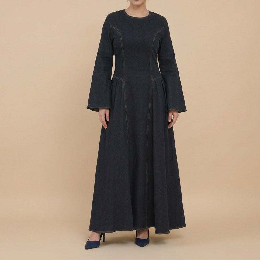 A woman modeling a long-sleeved, floor-length black dress with contrasting gray trim details on the sleeves, standing in a bright white studio setting with decorative pampas grass and a white chair.