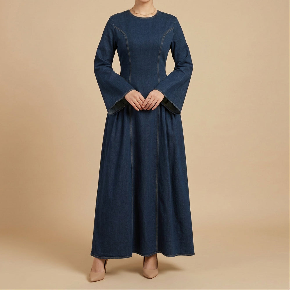 A woman wearing a long navy blue denim abaya dress with wide bell sleeves and contrast stitching, standing in a studio setting with a white background and decorative pampas grass.
