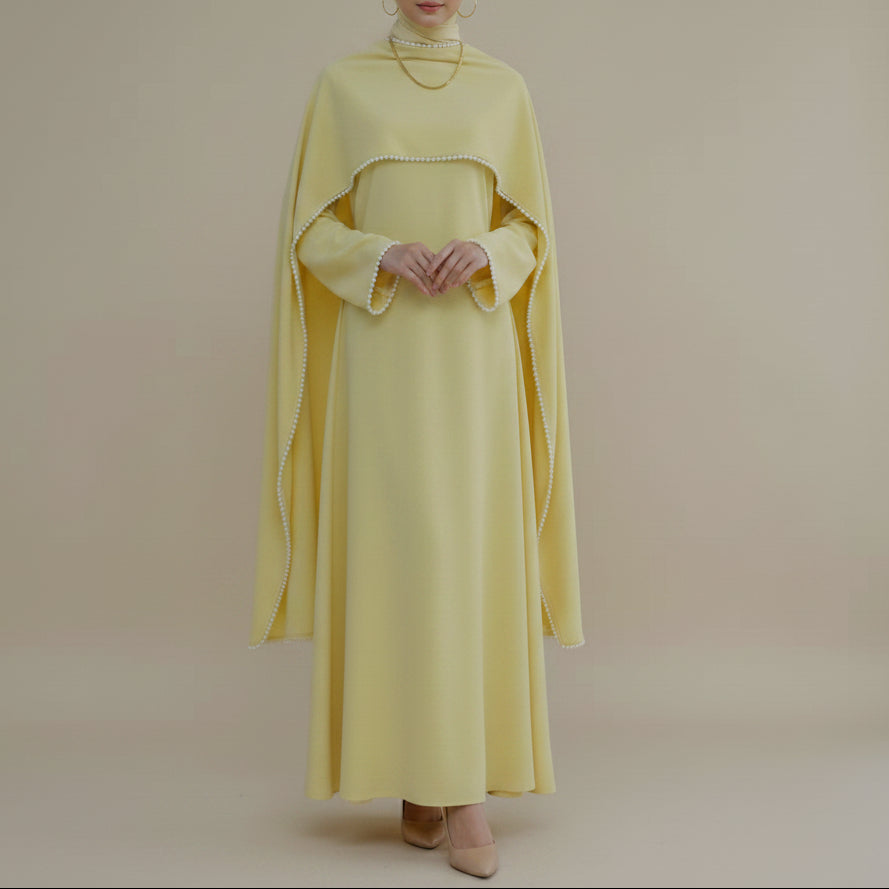 A pale yellow modest dress displayed on a mannequin in a bright, minimalist room. The dress features a draped shawl collar with delicate white trim and wide, flowing sleeves.