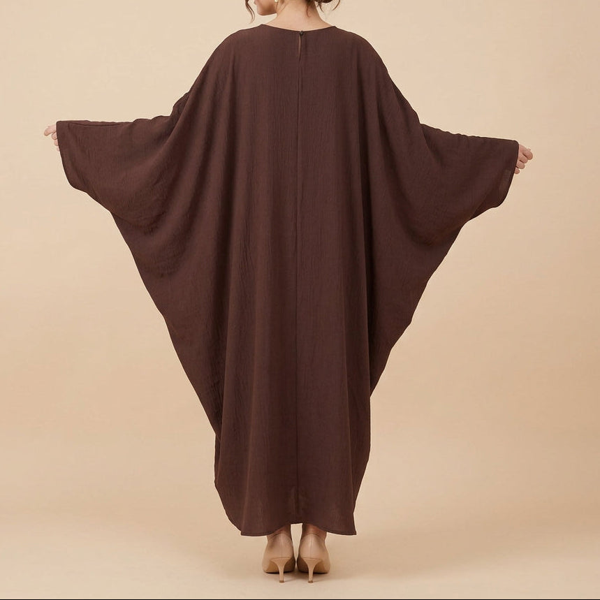 A woman viewed from behind wearing a loose, flowing brown caftan or tunic with very wide draped sleeves, standing against a plain white studio background.