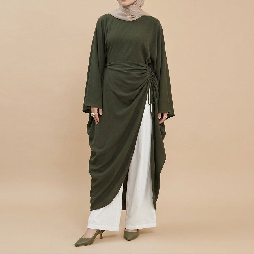 A model wearing an olive green long-sleeve draped maxi dress with white wide-leg pants and white sandals, photographed against a seamless white studio background.